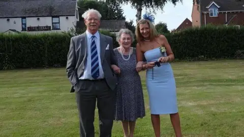 The Bays family David Bays pictured with his wife Janet and daughter Rachel at a wedding in 2017. The three are standing outside on a lawn, with the Five Bells pub in the background behind a hedge. David has white hair and a moustache and is wearing glasses. He is wearing a grey suit, white shirt and blue-striped tie. Janet is wearing a blue dress with white dots and has a long pearl necklace. Rachel is wearing a light blue strapless dress and blue fascinator, and is holding a camera.