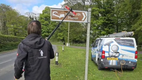 Linden Cleaning Services Jonny is stood in front of a brown sign for Hallgarth Manor. He is cleaning it with a red brush on a long pole. His Linden Cleaning Services van is parked to the right on grass.