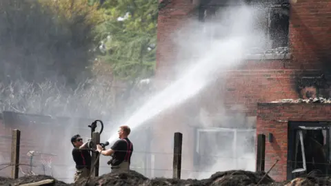 Reuters/Temilade Adelaja Two male firefighters operate a hose which blasts water at the winder of a house, with burn marks around the window.
