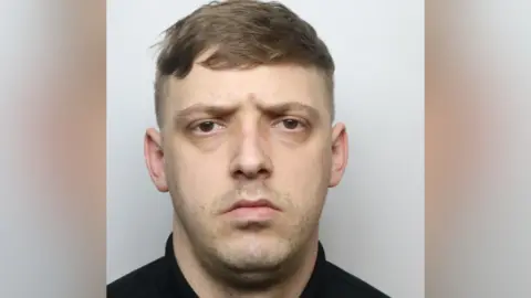 A man's police custody image. He has brown hair and short facial hair.