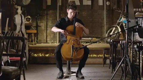 Dave Hills/Glude Media Sam Hills-Field playing a cello. He is wearing black with a stage behind him that has twinkling lights on it and the room is filled with wooden chairs.