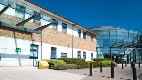 Google St George's Park Hospital exterior. There is a glass building on the right and a brick building on the left. Black bollards are installed in the paved area.