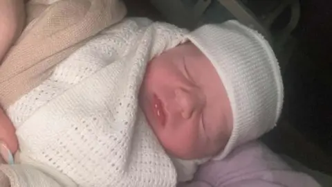 The baby is wearing a white hat and is wrapped in a white blanket. His eyes are closed. He is being held by someone with blue and pink nails.