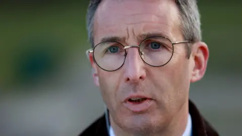 Andrew Muir is speaking to the press wearing a brown jacket and has glasses on. He has short white/grey hair.