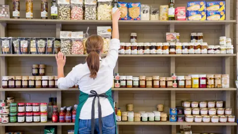 An anonymous woman wearing an apron, photographed from behind, stocking a supermarket shelf