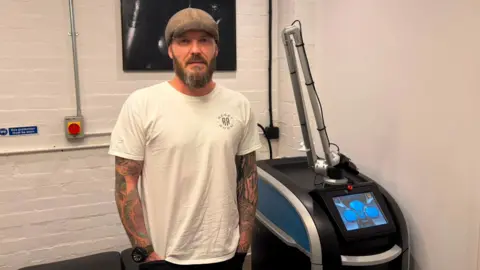 A heavily tattooed man with a white and grey beard wearing a white t-shirt, a flat cap is standing in a white room next to a laser machine.