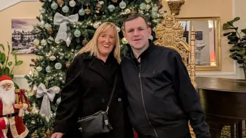 David Green The photo shows a man and a woman smiling at the camera. The woman on the left has shoulder length blonde hair and is wearing a black coat with a black bag across her body. The man on the right is also wearing a black jacket. They're standing in front of a Christmas tree. 
