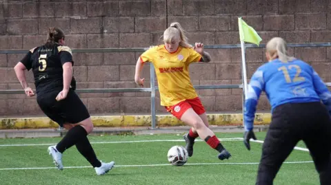 JazzaMcG Photography A Stewarton Annick Ladies FC player on the ball, wearing the club kit of yellow short and red shorts. She is is taking on a defender on the other team, wearing an all black kit, and watched by the other team's goalkeeper, wearing a blue shirt and black jogging bottoms.