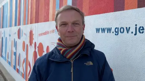 BBC A man wearing a navy coat with a stripy scarfed tucked in standing in front of the climate change mural.