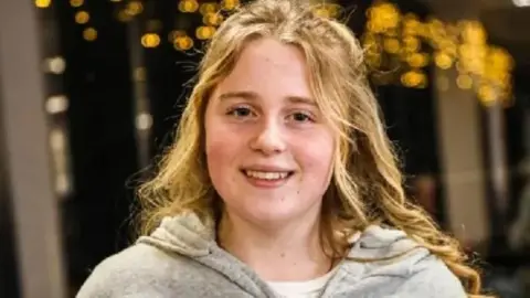 Chloe pictured wearing grey hoodie and white t-shirt and smiling. She has long wavy blonde hair