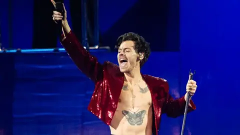Getty Images Harry Styles is wearing an open sparkly red jacket. He's on stage singer with one hand in the air holding a mic, his hair is brown and curly. He has a tattoo of a butterfly on his torso. 