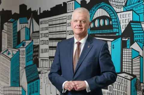 University of Sunderland A man with close-cropped white hair, wearing a suit, holds his hands in front of an illustrated mural of Sunderland