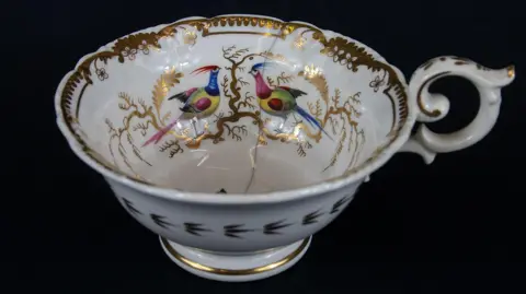 An ornate and dainty teacup from the 19th Century, which is decorated in a gold filigree pattern and features a motif of two blue and green peacocks facing each other on the inside of the cup.