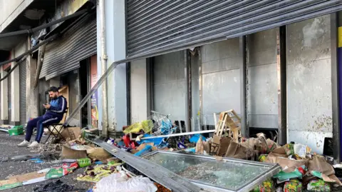 PA Media: Front of a burnt out shop and the owner sitting on a chair in the street