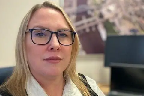 Gateshead's Director of Public Health, Alice Wiseman. She has shoulder-length blonde hair and is wearing black-rimmed glasses and a white shirt. She is pictured at her desk, in front of an aerial photograph of Gateshead