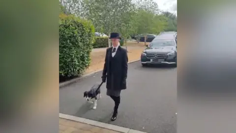A woman in a black suit with a white shirt, black shoes, gloves and black bowler hat is walking in front of a hearse with a dog 