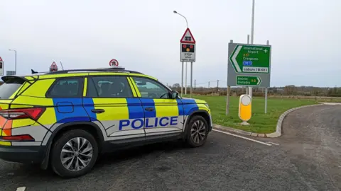 A police car is blockimg a road, close to a sign that shows a railway crossing is ahead. 