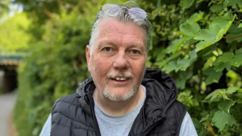 A man wearing a t-shirt and a puffer vest stands smiling in front of a green leafy bush. 