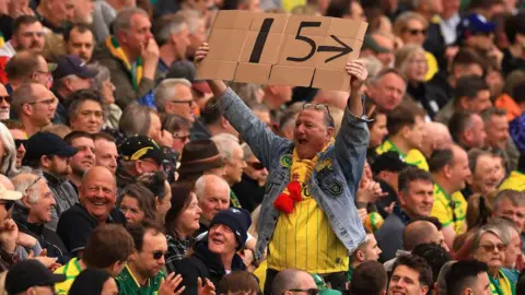Getty Images In a stand packed with fans who are seated, one fan is standing up. He is facing away from the pitch and is wearing a blue denim jacket over a yellow Norwich City shirt. He has his glasses on his head. He is cheering and holding a large cardboard sign over his head which reads "15", the number of years Norwich have gone undefeated in the derby.