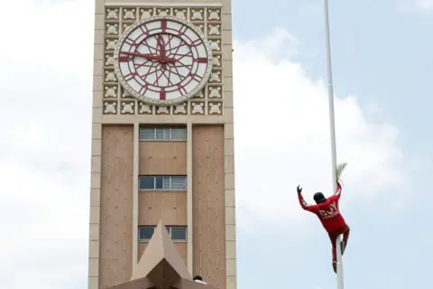 MONICAH MWANGI / REUTERS A man climbs up a tall pole with the parliament tower seen behind it.