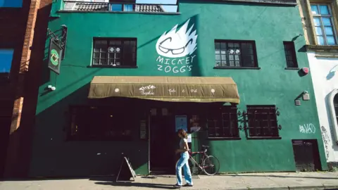 A woman walking outside of a green pub called Mickey Zoggs
