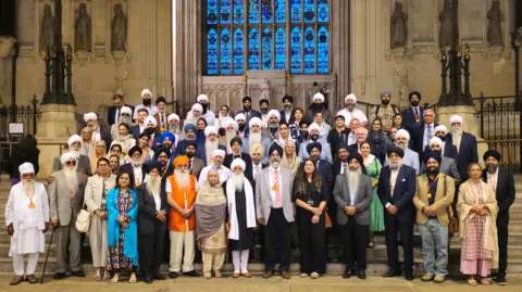 Nishkam Media A group of Sikhs standing on steps in Parliament. Behind them is a large stain glass window.