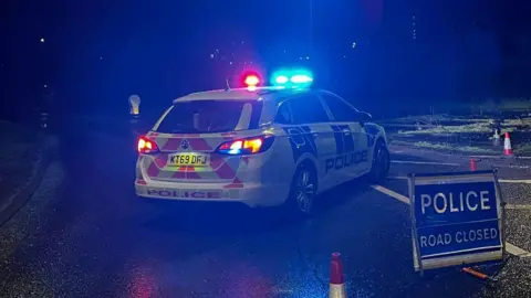 A police car parked at an angle across a sliproad blocks the entrance to a road. A "POLICE ROAD CLOSED" sign is positioned in the foreground.