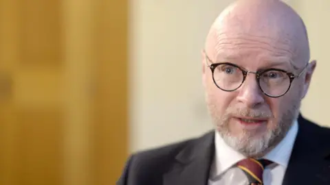 Liam Byrne has a greyish beard and wears a suit and glasses. He sits to the right of the camera and is photographed as he is being interviewed. 