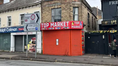 A shop with a bright orange closed shutter and sign reading Top Market between a pub and another shop with a road sign reading Minehead and town centre on the pavement outside