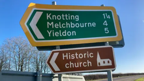 Alex Pope/BBC Two road signs with different names on them, one brown saying Historic Church. There is a road behind the signs and trees in the distance. The sky is blue. 