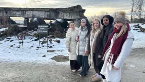 Five young women aged 18 stand solemnly beside a ruined crematorium at Auschwitz Birkenau