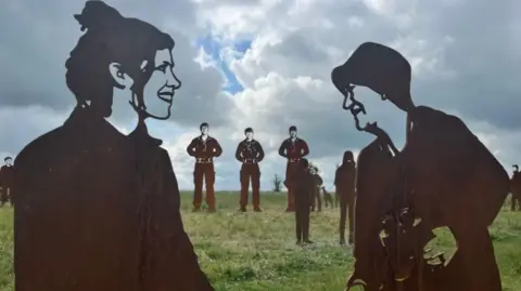 Statues of military personnel on military grounds in Salisbury. In the center there are two female figures staring at each other. On the left, the statue of a woman with her hair up and smiling looking at a female figure on the right. The figure on the right is holding a pair of binoculars. 