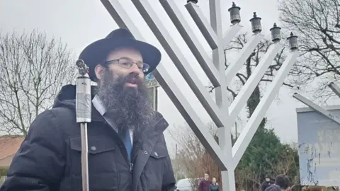 Rabbi Eliezer Tunk stands next to a menorah, a wooden structure with eight branches, at a lighting ceremony. There are people in the background. The damaged menorah, which has branches broken off, is also in the background.