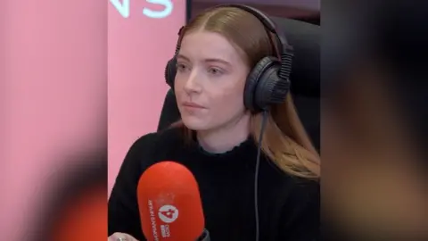 A woman with long brown hair wearing a black top sits in a radio studio in front of a microphone