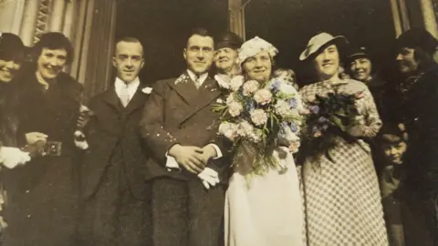 Beryl Qualtrough A sepia toned photo of a wedding a bride and groom surrounded by the rest of the wedding party.
