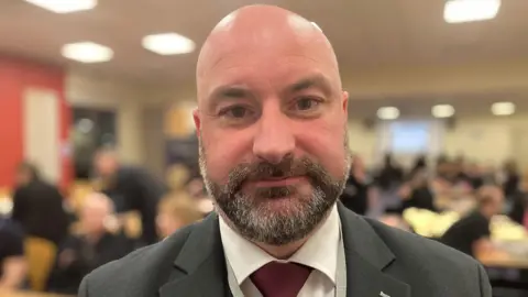 Sharon Edwards/BBC Head and shoulders shot of Marc Jones. He is wearing a grey suit and red tie and is looking directly at the camera. The background of the image is blurred but shows people sitting and standing at desks.