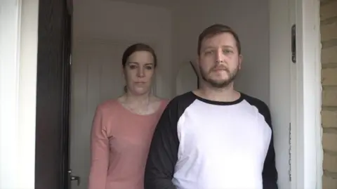 A man wearing a black and white t-shirt and a woman wearing a pink t-shirt stood in a doorway. They look cross.