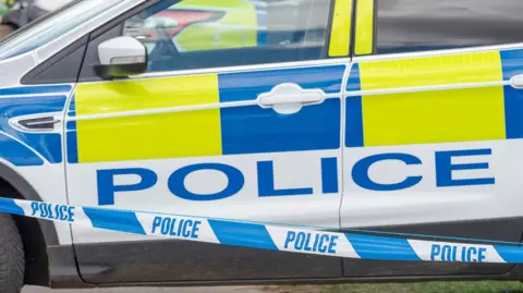 A stock image showing a parked police car with hi-vis blue and yellow sticker decals. In the foreground there is blue and white police cordon tape.