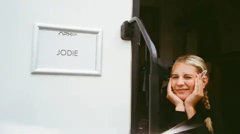Callie Cooke/Instagram Callie Cooke looking at the camera and squinting. She is in the doorway of her trailer with her head in her cupped hands. To her right is a sign saying "Jodie".