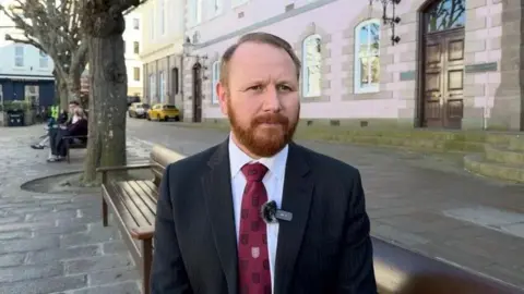 Tom Coles is standing in the street being interviewed and has a small fluffy microphone on his lapel. He has short light-coloured hair and a ginger moustache and beard. He is wearing a white shirt, burgundy patterned tie and black pin-striped jacket. Behind him are two wooden benches on the cobbled street and there are tress next to them. The surrounding buildings are pastel-coloured. 