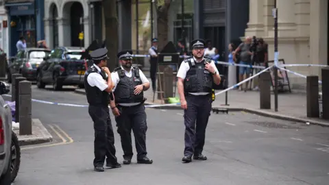 PA Media Three police officers are pictured standing on a street in front a police cordon. 