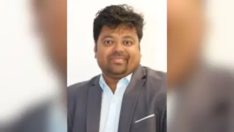 Portrait image of Leedo George. He is looking into camera and has dark hair, a short goatee beard and is wearing a suit jacket and shirt with the top button undone.