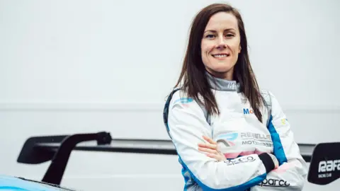 Racing driver Abbie Eaton stands next to the rear spoiler of her car. She has shoulder-length brown hair and wears a white and blue race suit. 