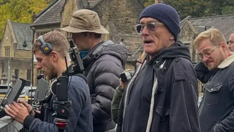 Willow Tree Cafe at No 5 Image shows a group of five or six men all wearing dark coats. The men to the left of the picture are holding cameras and wearing headphones. The man in the centre - Danny Boyle - is wearing a dark jacket, a hat and sunglasses. 