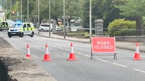 Police vehicles, police tape and police officers at scene of crash bike on road, in between two parks, with a road closed sign.