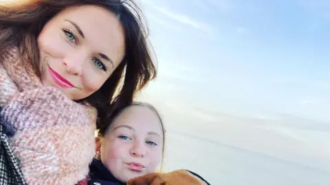 A woman with dark hair and wearing a checked coat and a pink scarf smiles at the camera, while next to her is a young girl with blonde hair and, just seen, the top of a dog's head