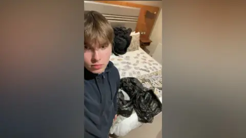 Metropolitan Police A selfie image of a teenage boy dressed in a black zip up top. He has light brown hair with a long fringe. Behind him s a bed with cash notes laid out and a black bin liner next to them.