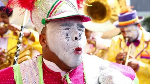 A man wearing white face paint with a colourful elabourate hat and pink outfit. His eyes are closed and he is making an expressive expression.