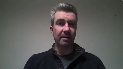 A headshot of Donnelly, who has short grey hair. He is wearing a dark coloured fleece and a grey top underneath. He is looking directly at the camera. 