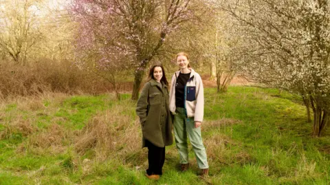 BBC Two women stood in front of a blossom tree. The woman on the right has red hair with a cream jacket and black t-shirt with light green trousers. The woman on the left has long brown hair and is wearing a green full-length coat with black trousers